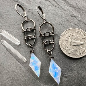 May include: A pair of dangle earrings featuring dark metal lever-back hooks, three interconnected circular links, and a diamond-shaped moonstone pendant with blue iridescence. Two clear quartz points and a US quarter are shown for scale.