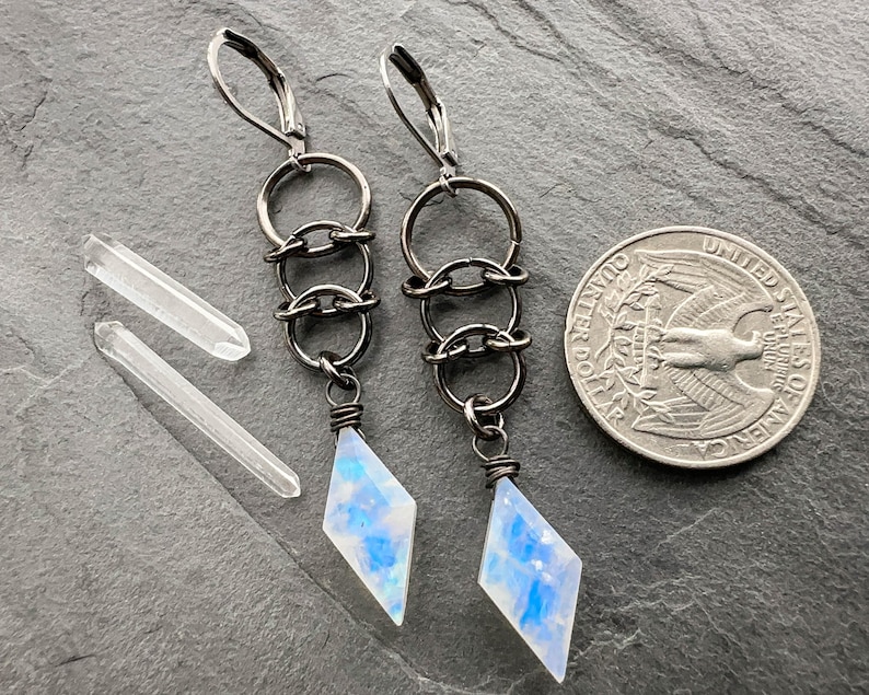 May include: A pair of dangle earrings featuring dark metal lever-back hooks, three interconnected circular links, and a diamond-shaped moonstone pendant with blue iridescence. Two clear quartz points and a US quarter are shown for scale.