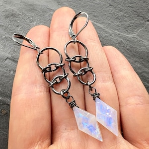 May include: A pair of dangle earrings featuring dark gunmetal-toned wire loops connected in a vertical chain. Each earring terminates in a translucent, diamond-shaped stone with iridescent blue flashes, secured by a wire-wrapped setting.