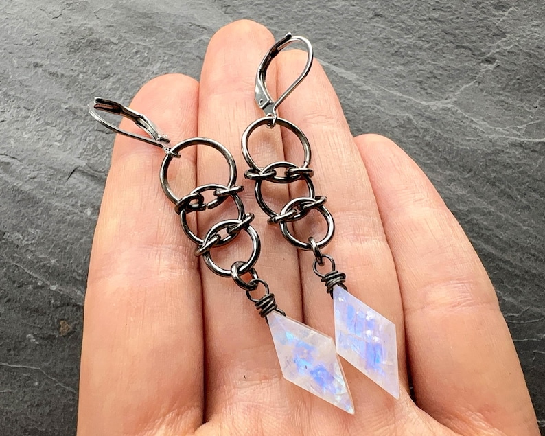 May include: A pair of dangle earrings featuring dark gunmetal-toned wire loops connected in a vertical chain. Each earring terminates in a translucent, diamond-shaped stone with iridescent blue flashes, secured by a wire-wrapped setting.