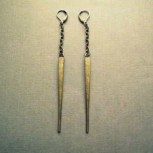 Pu&ograve; includere: Un paio di orecchini pendenti lunghi e sottili in ottone anticato. Ogni orecchino presenta una chiusura a monachella collegata a una corta catena metallica, che sostiene un lungo pendente a punta affusolato e testurizzato.