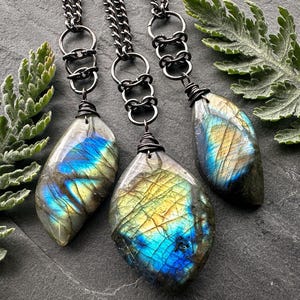 May include: Three necklaces with labradorite gemstone pendants. The pendants are teardrop shaped and have a blue, green, and gold iridescent sheen. The necklaces are made with black metal chain and wire.