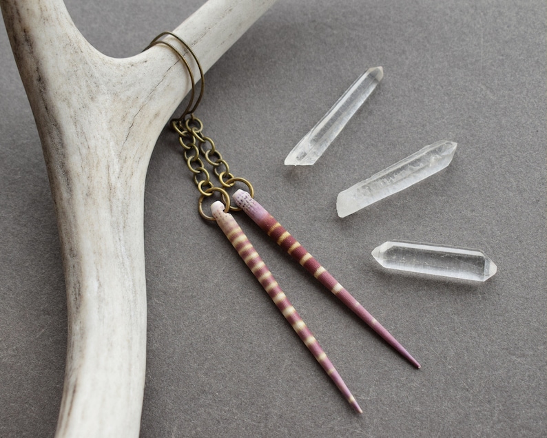 May include: Two long, thin, pink and white striped earrings with a brass chain and hook. The earrings are hanging from a white antler on a gray background.