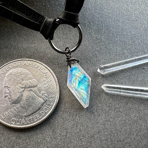 K&ouml;nnte beinhalten: Eine silberne Halskette mit einem schwarzen Lederband und einem schillernden blau-gr&uuml;nen Kristallanh&auml;nger. Der Anh&auml;nger ist mit einem silbernen Draht umwickelt am Band befestigt. Zwei klare Quarzkristallspitzen sind ebenfalls enthalten.