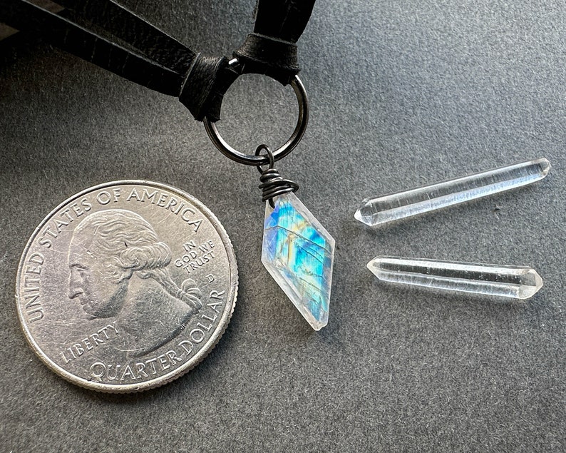 K&ouml;nnte beinhalten: Eine silberne Halskette mit einem schwarzen Lederband und einem schillernden blau-gr&uuml;nen Kristallanh&auml;nger. Der Anh&auml;nger ist mit einem silbernen Draht umwickelt am Band befestigt. Zwei klare Quarzkristallspitzen sind ebenfalls enthalten.
