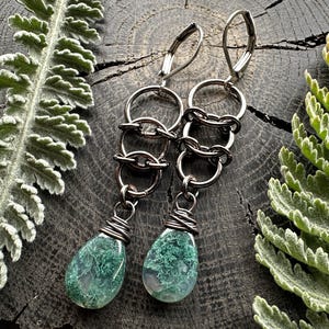 May include: A pair of dangle earrings featuring dark metallic lever-back hooks connected to three interlocking circular rings. Each earring terminates in a teardrop-shaped green moss agate gemstone wrapped at the top with dark wire.
