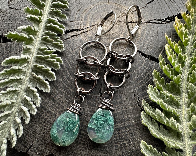 May include: A pair of dangle earrings featuring dark metallic lever-back hooks connected to three interlocking circular rings. Each earring terminates in a teardrop-shaped green moss agate gemstone wrapped at the top with dark wire.