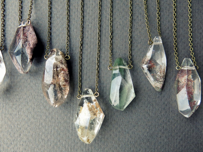 May include: A collection of eight crystal pendants on delicate chains. The crystals are clear with inclusions of brown, green, and red. The pendants are all different shapes and sizes.