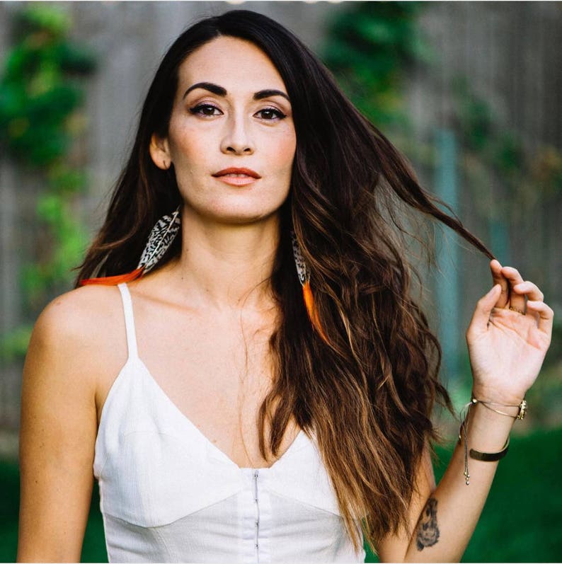 May include: A woman with long brown hair wearing a white tank top and feather earrings. She is holding a strand of her hair in her right hand.