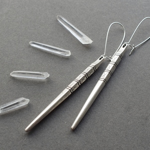 May include: A pair of silver earrings with a geometric design. The earrings are long and dangle, and they have a textured surface.