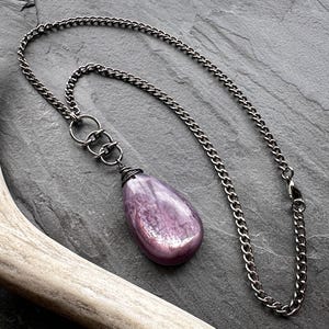 K&ouml;nnte beinhalten: Eine silberfarbene Halskette mit einem gro&szlig;en, tropfenf&ouml;rmigen, lila Edelsteinanh&auml;nger. Der Anh&auml;nger ist mit Draht umwickelt und mit drei kleinen Ringen an der Kette befestigt. Die Halskette liegt auf einer dunkelgrauen, strukturierten Oberfl&auml;che.