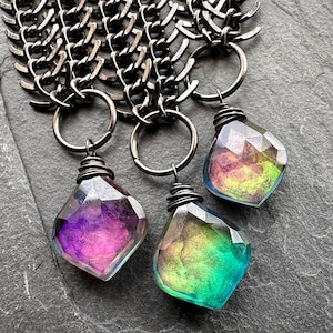 May include: Three faceted pendants with iridescent color gradients of purple, green, and yellow are suspended from a silver chain. Each pendant is wire-wrapped and attached to a silver ring. The chain is made of interlocking links.