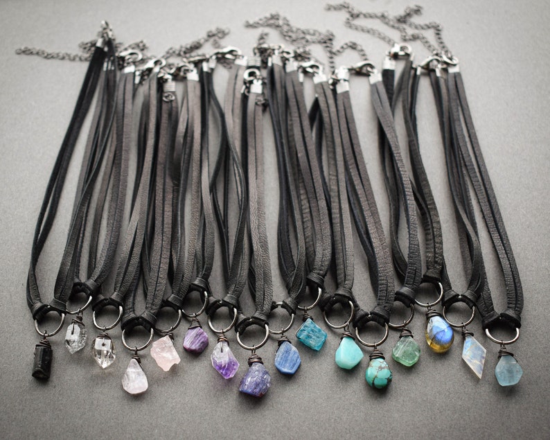 K&ouml;nnte beinhalten: Eine Sammlung von 15 Lederhalsketten mit verschiedenen Edelsteinanh&auml;ngern. Die Halsketten sind schwarz und die Anh&auml;nger sind in verschiedenen Farben, darunter klar, rosa, lila, blau und gr&uuml;n.