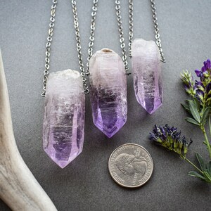 May include: Three silver chains with amethyst crystal pendants. The crystals are pointed and have a light purple color. The chains are hanging against a gray background.