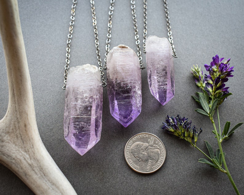 May include: Three silver chains with amethyst crystal pendants. The crystals are pointed and have a light purple color. The chains are hanging against a gray background.