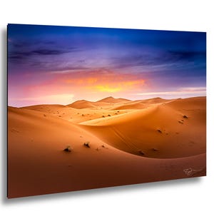 May include: A landscape photograph of golden sand dunes under a vibrant sunset sky. The horizon features rolling desert hills with soft shadows, while the sky transitions from deep blue to purple and bright orange near the sun's glow.