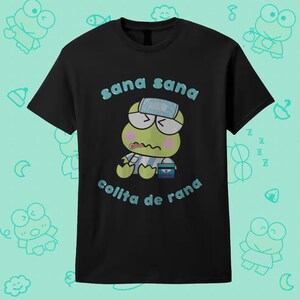 May include: A black short-sleeved t-shirt featuring a graphic of a green frog character wearing a blue and white striped shirt and a head compress. The frog has a thermometer in its mouth. The text "sana sana colita de rana" is printed in blue.