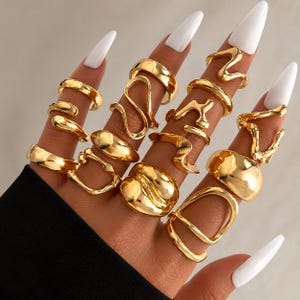 May include: A collection of gold-toned rings worn on fingers, featuring various designs including smooth bands, open-ended shapes, wavy lines, and chunky rounded forms. The rings have a polished, reflective finish and are worn across multiple fingers.