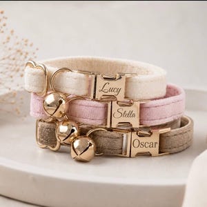 May include: Three pet collars in cream, light pink, and beige velvet fabric. Each collar features a gold-toned metal side-release buckle engraved with the names Lucy, Stella, and Oscar, respectively. A gold-toned bell is attached to each collar.