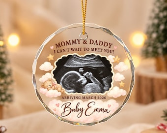 Può includere: Un ornamento rotondo in acrilico trasparente con un'ecografia al centro, circondato da nuvole rosa, stelle dorate e un orsetto. Il testo recita: MOMMY & DADDY I CAN'T WAIT TO MEET YOU!, ARRIVING MARCH 2026, e il nome Baby Emma.
