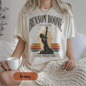 May include: An ivory short-sleeve t-shirt featuring a graphic print of a singer kneeling with a microphone. The text reads BENSON BOONE WANTED MAN and 2026 WORLD TOUR. The design includes a sunburst background and horizontal stripes in muted colours.
