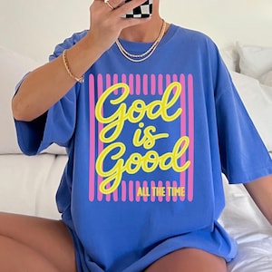 May include: A periwinkle blue short-sleeve t-shirt featuring a graphic design with vertical pink stripes. The text "God is Good" is printed in a large yellow cursive font, with the words "ALL THE TIME" in a smaller yellow block font below.