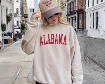 May include: A beige crewneck sweatshirt featuring the word ALABAMA in large red capital letters across the chest. The outfit includes a beige and grey baseball cap with the text The Western House Casino Las Vegas Nevada printed on the front.