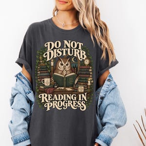 May include: A charcoal gray t-shirt featuring a graphic of an owl reading a book with the text DO NOT DISTURB READING IN PROGRESS. The design includes stacks of books, a candle, a mug, and ivy leaves surrounding the central owl illustration.