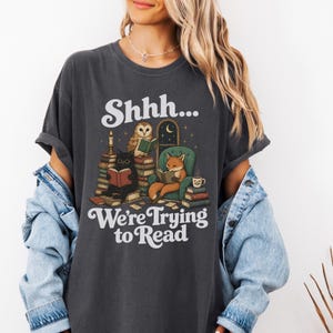 Può includere: Una t-shirt grigio antracite con un'illustrazione grafica di un gufo, un gatto nero e una volpe che leggono libri. Il testo "Shhh... We're Trying to Read" è scritto in bianco. Il disegno include pile di libri, una candela e una poltrona.