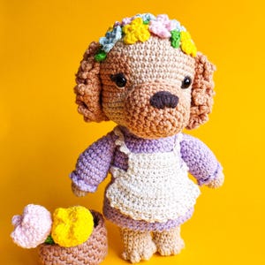 Peut inclure: Une poupée chien au crochet avec une fourrure beige, des yeux noirs et un nez foncé, portant une robe lavande avec un tablier blanc et une couronne de fleurs colorées. À côté de la poupée se trouve un petit panier beige contenant deux fleurs.