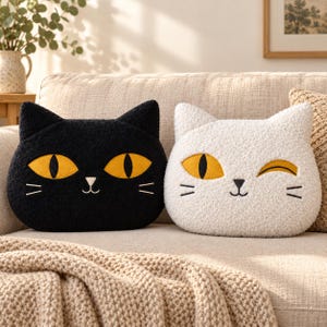Pode incluir: Duas almofadas em forma de gato feitas de tecido texturizado. Uma almofada é preta com dois olhos amarelos abertos, enquanto a outra é branca com um olho amarelo aberto e um olho amarelo a piscar. Ambas têm narizes e bigodes pretos bordados.