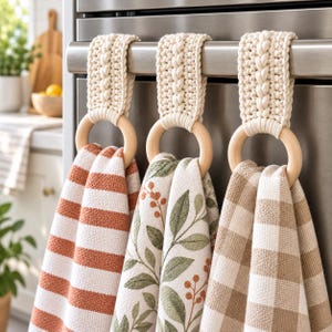 Op de afbeelding: Drie keukendoeken hangen aan een roestvrijstalen ovenhandgreep. Elke doek is bevestigd met een crèmekleurige gevlochten macraméband aan een gladde houten ring. De doeken hebben oranje strepen, groene bladpatronen en een beige ruitpatroon.