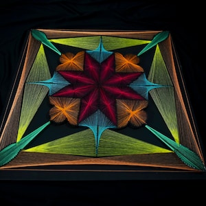 May include: A square string art piece on a black base featuring a central eight-pointed star in deep red, surrounded by orange, light blue, and lime green geometric patterns created with taut threads arranged in symmetrical, radiating lines.