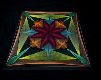 May include: A square string art piece on a black base featuring a central eight-pointed star in deep red, surrounded by orange, light blue, and lime green geometric patterns created with taut threads arranged in symmetrical, radiating lines.
