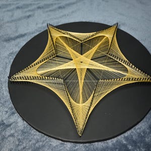 May include: A circular black board featuring a star-shaped string art design created with thin, light-coloured thread wrapped around numerous small black nails. The geometric pattern forms a central pentagon with radiating lines extending to the points.