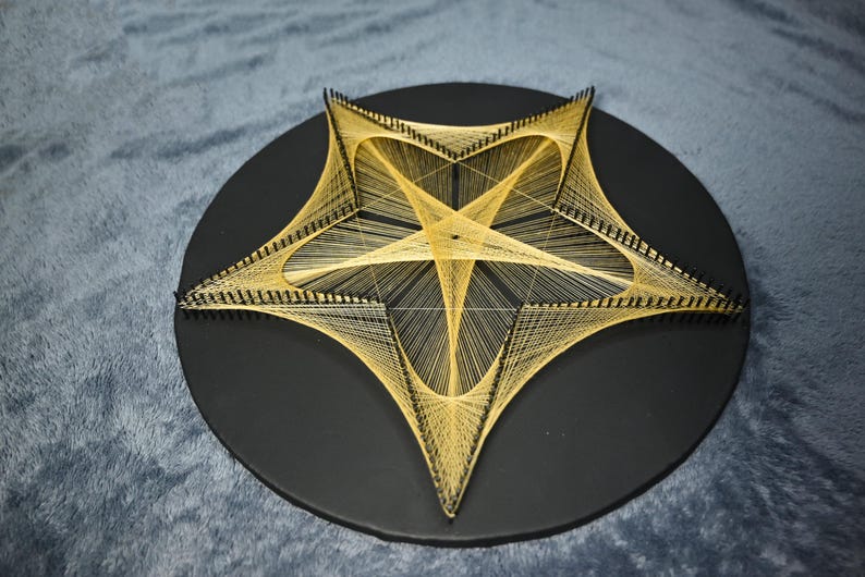 May include: A circular black board featuring a star-shaped string art design created with thin, light-coloured thread wrapped around numerous small black nails. The geometric pattern forms a central pentagon with radiating lines extending to the points.