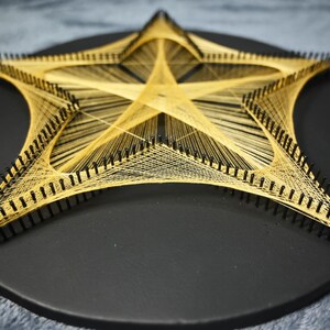 May include: A star-shaped string art piece featuring gold-coloured thread woven between black nails arranged in a star pattern on a circular black wooden base. The intricate geometric design creates a central star shape with radiating lines.