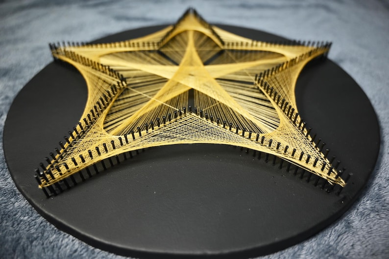 May include: A star-shaped string art piece featuring gold-coloured thread woven between black nails arranged in a star pattern on a circular black wooden base. The intricate geometric design creates a central star shape with radiating lines.
