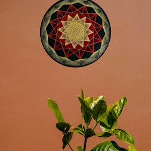 May include: A circular string art piece featuring a black background with a geometric star pattern created from yellow and red thread. The design consists of a central yellow star surrounded by layers of red and yellow radiating lines.