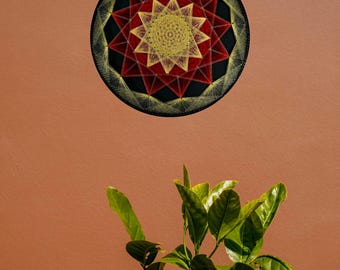 May include: A circular string art piece featuring a black background with a geometric star pattern created from yellow and red thread. The design consists of a central yellow star surrounded by layers of red and yellow radiating lines.