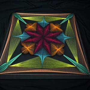 May include: A square string art piece on a black base featuring a central eight-pointed star in magenta, surrounded by orange, light blue, and lime green geometric patterns created with taut threads arranged in symmetrical, radiating lines.