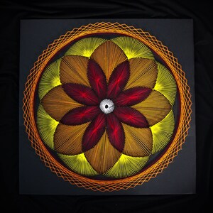 May include: A square string art piece featuring a circular mandala design. The pattern consists of layered threads in shades of bright yellow, orange, deep red, and brown, radiating from a white centre point, enclosed by an orange geometric border.