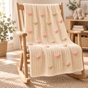 Puede incluir: Una manta de punto color crema con una textura acanalada vertical y aplicaciones de corazones tridimensionales dispersos en tonos suaves de rosa y beige. La manta está colocada sobre una mecedora de madera clara con respaldo de listones.