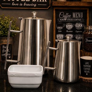 May include: Two stainless steel coffee carafes with lids and angular handles, positioned behind a small, rectangular white ceramic bowl. The carafes have a polished metallic finish and a tapered cylindrical shape, standing on a dark surface.