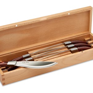 May include: A set of four steak knives with dark wood handles and silver metal bolsters, presented in a rectangular light-colored wooden storage box. One knife rests outside the box, while three are stored inside, featuring engraved branding.