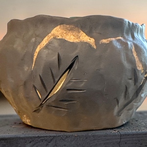Może przedstawiać: Ciemnoszara, ręcznie formowana miska ceramiczna o nierównej, teksturowanej powierzchni. Zewnętrzna strona posiada wycięte wzory liści oraz złote metaliczne akcenty na górnej krawędzi i w zagłębieniach wzorów liści.