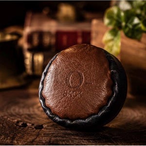 May include: A circular, two-toned object featuring a textured brown leather-like center panel surrounded by a smooth black border. The center panel is embossed with the letter O and the word BACK positioned directly underneath the letter.
