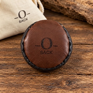 May include: A round footbag made of brown leather with a black leather border. The center of the brown leather features the black text -O- SACK. A matching beige fabric drawstring pouch with the same -O- SACK logo is positioned behind the footbag.