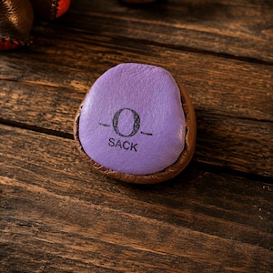 May include: A round, multi-colored footbag or hacky sack featuring a purple leather top panel with the black printed text O SACK. The sides are constructed from brown leather, and the object is stitched together with visible dark thread seams.