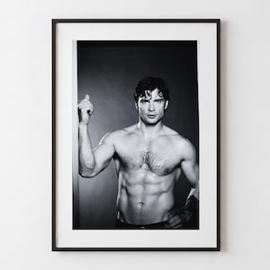Op de afbeelding: Een zwart-wit fotoprint in een dunne zwarte lijst met een persoon zonder shirt, donker haar, een gedefinieerde romp en borsthaar, vastgelegd vanaf de taille tegen een donkere achtergrond met één arm lichtjes omhoog geheven.